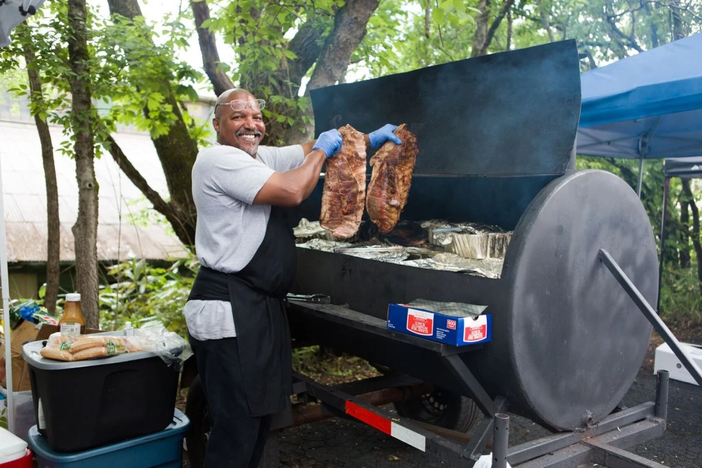 a smiling man holds up meat above a barbecue grill